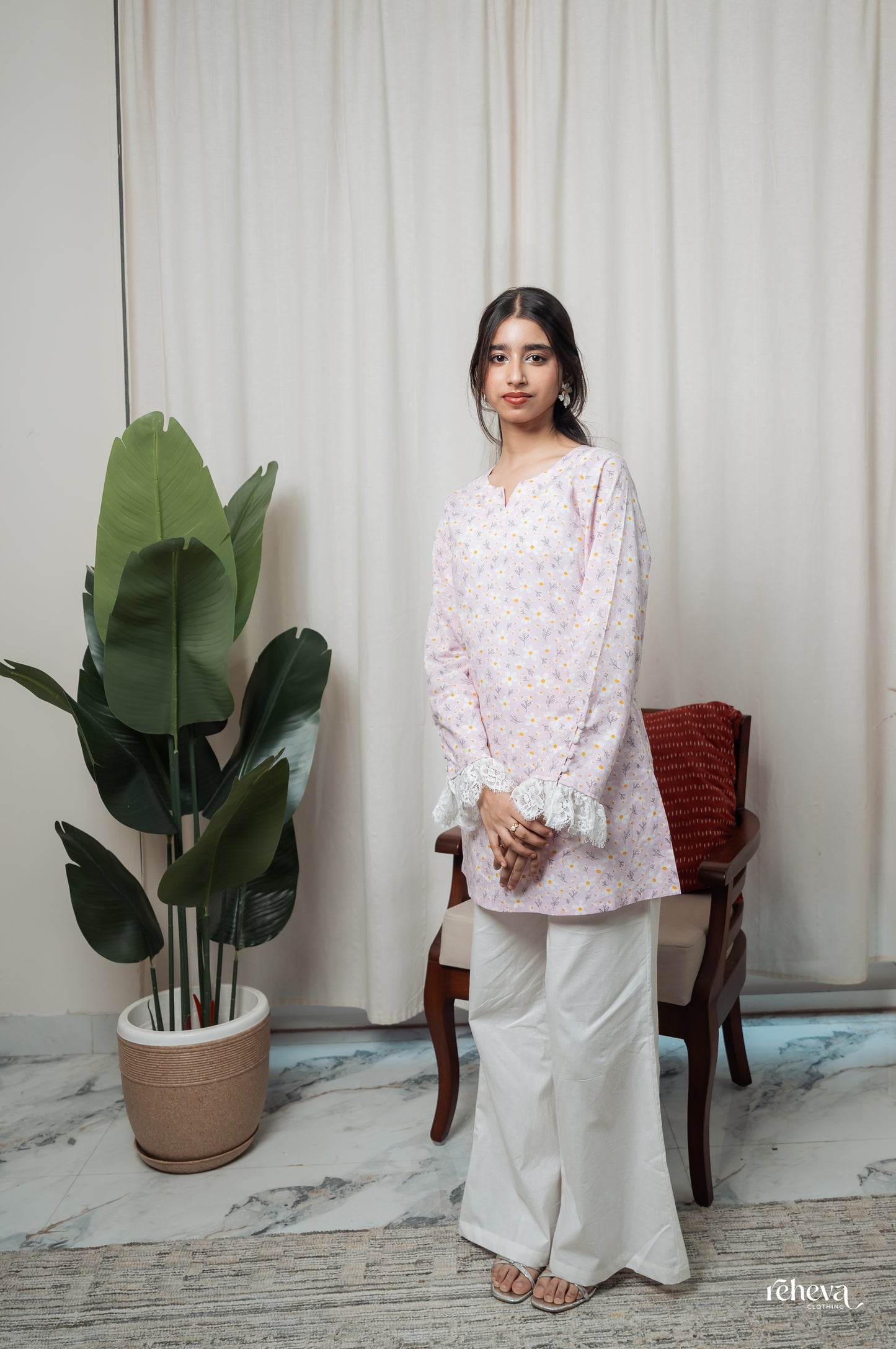 A woman stands indoors wearing a light lavender long-sleeve kurta with a small multicolor floral print and white wide-leg pants. She is posed next to a potted plant and a wooden chair with a red cushion, in front of cream-colored curtains.