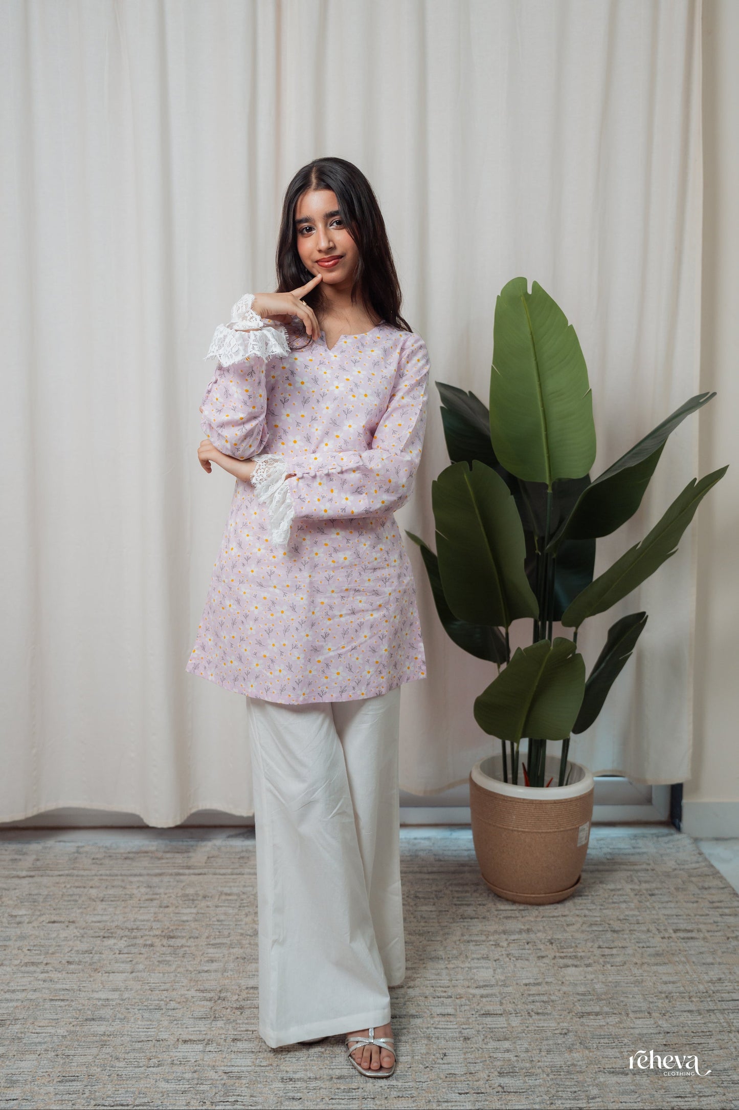 A woman modeling a light lavender floral printed kurta with white wide-leg pants, standing in front of a cream curtain next to a large green potted plant.