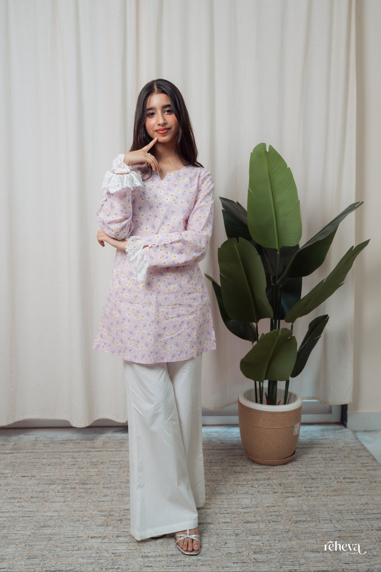 A woman modeling a light lavender floral long-sleeve tunic top with white wide-leg pants, standing in front of a beige curtain next to a potted green plant.