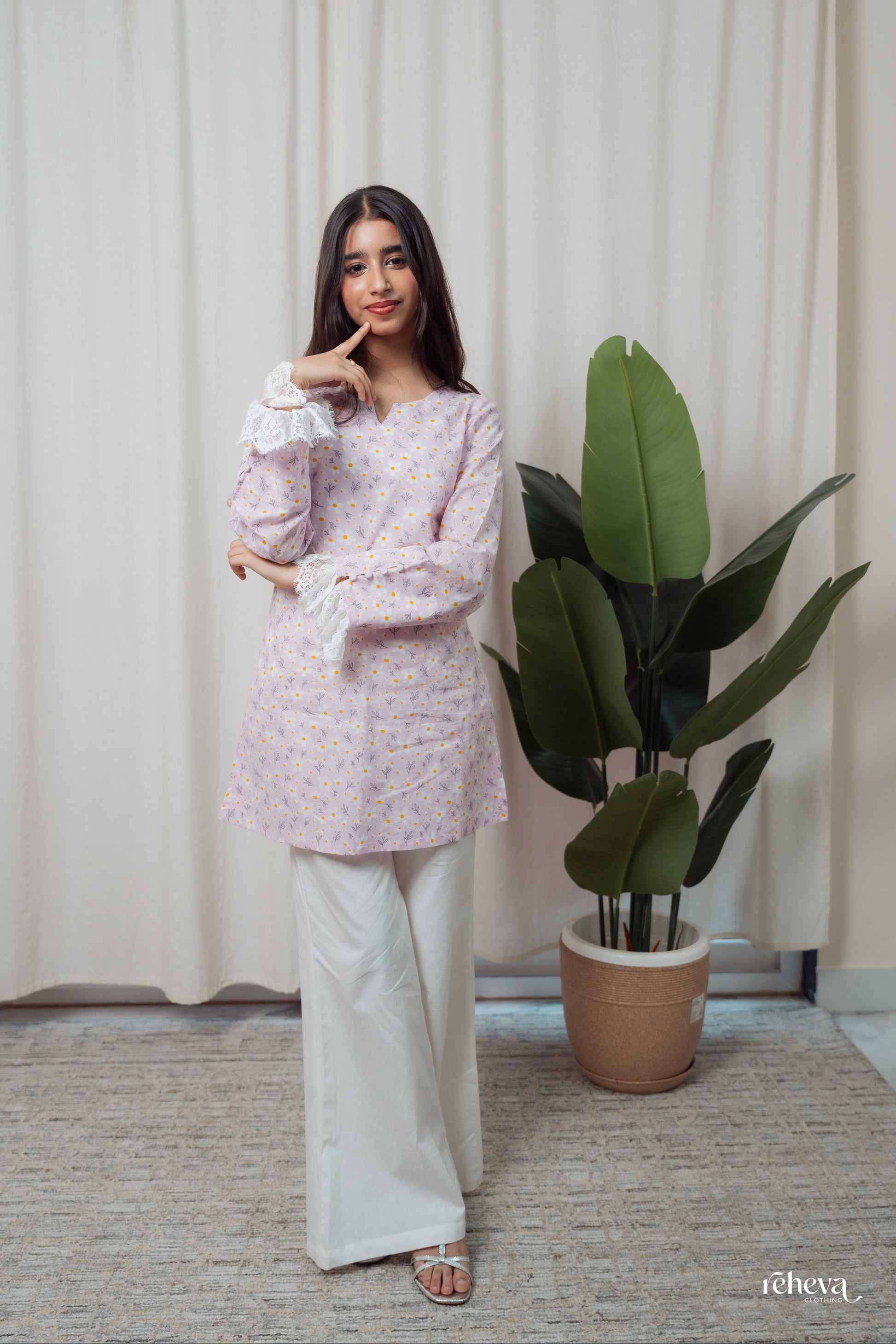 A woman modeling a light lavender floral long-sleeve tunic top with white wide-leg pants, standing in front of a beige curtain next to a potted green plant.