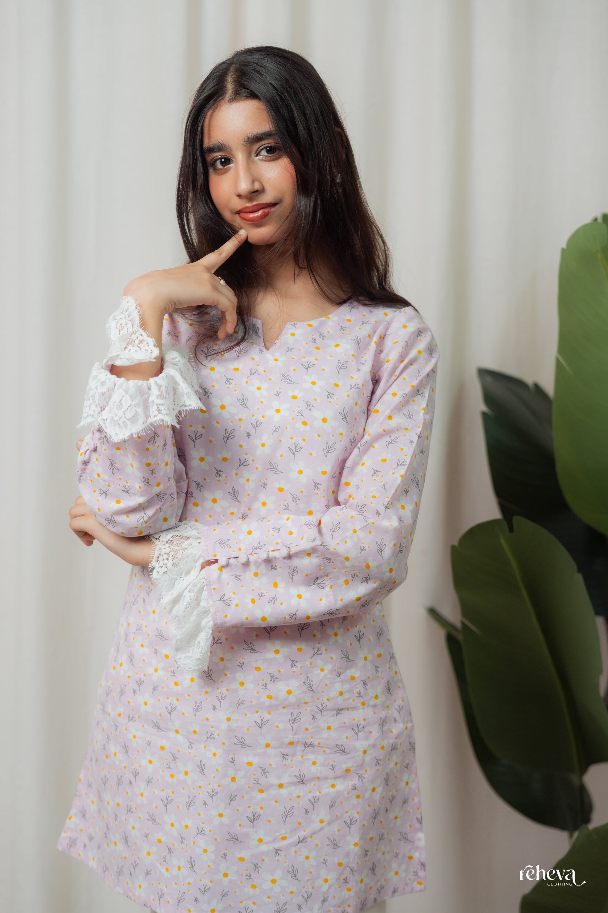 A young woman modeling a light lavender long-sleeve tunic dress with a small multicolor floral print, white lace cuffs, and a V-neckline, posed against a neutral cream curtain backdrop with a green plant leaf visible on the right.