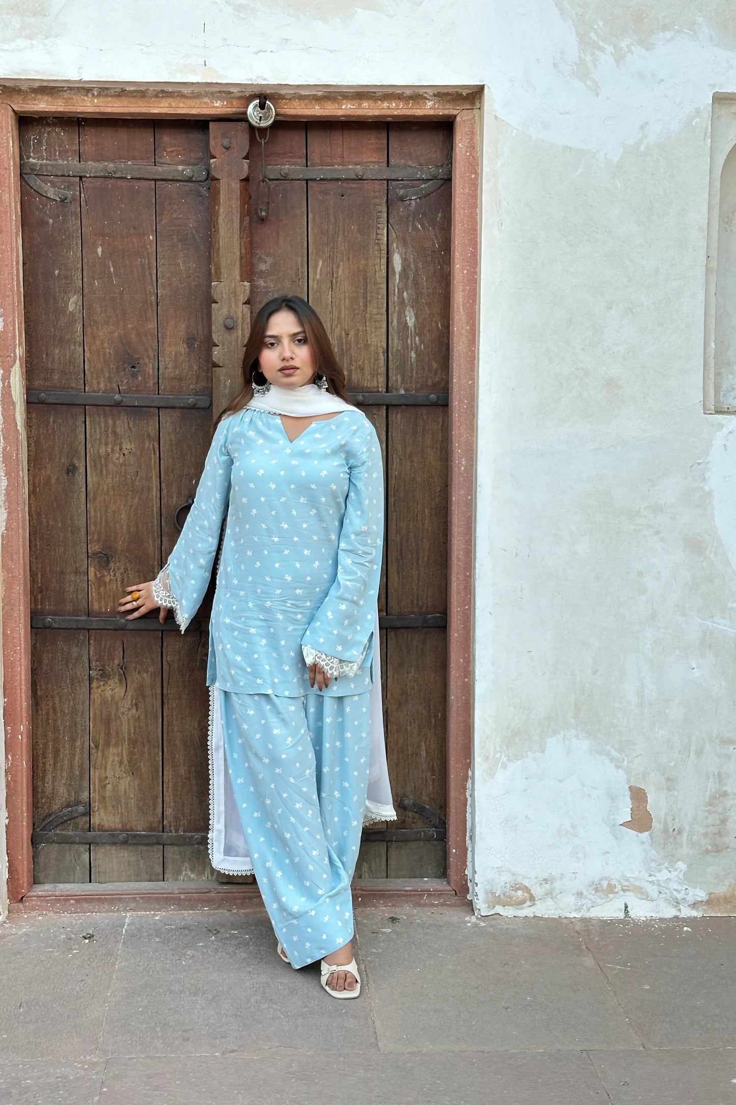 A woman wearing a light blue and white patterned traditional outfit stands in front of a rustic wooden door and textured wall.