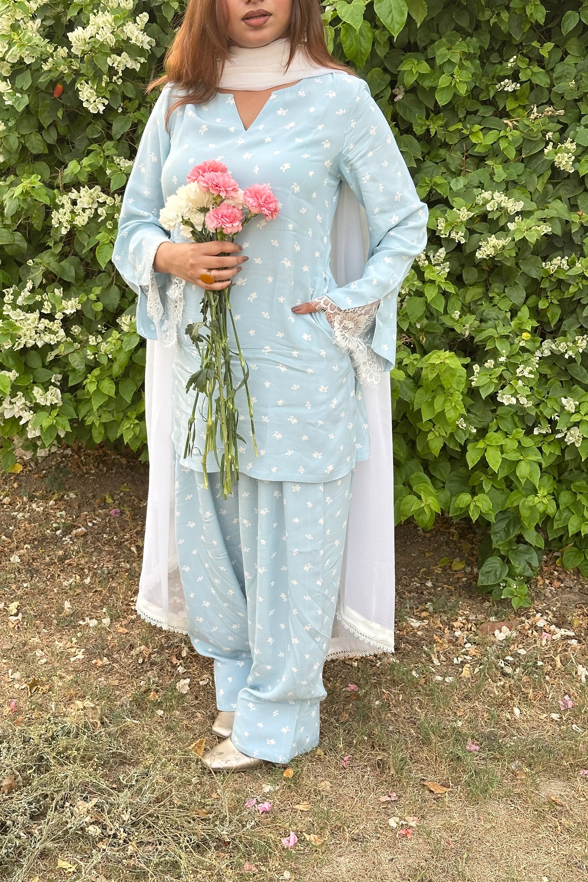 A woman wearing a light blue three-piece salwar kameez set with white floral prints, standing outdoors in front of a green leafy hedge and holding a small bouquet of pink and white flowers.