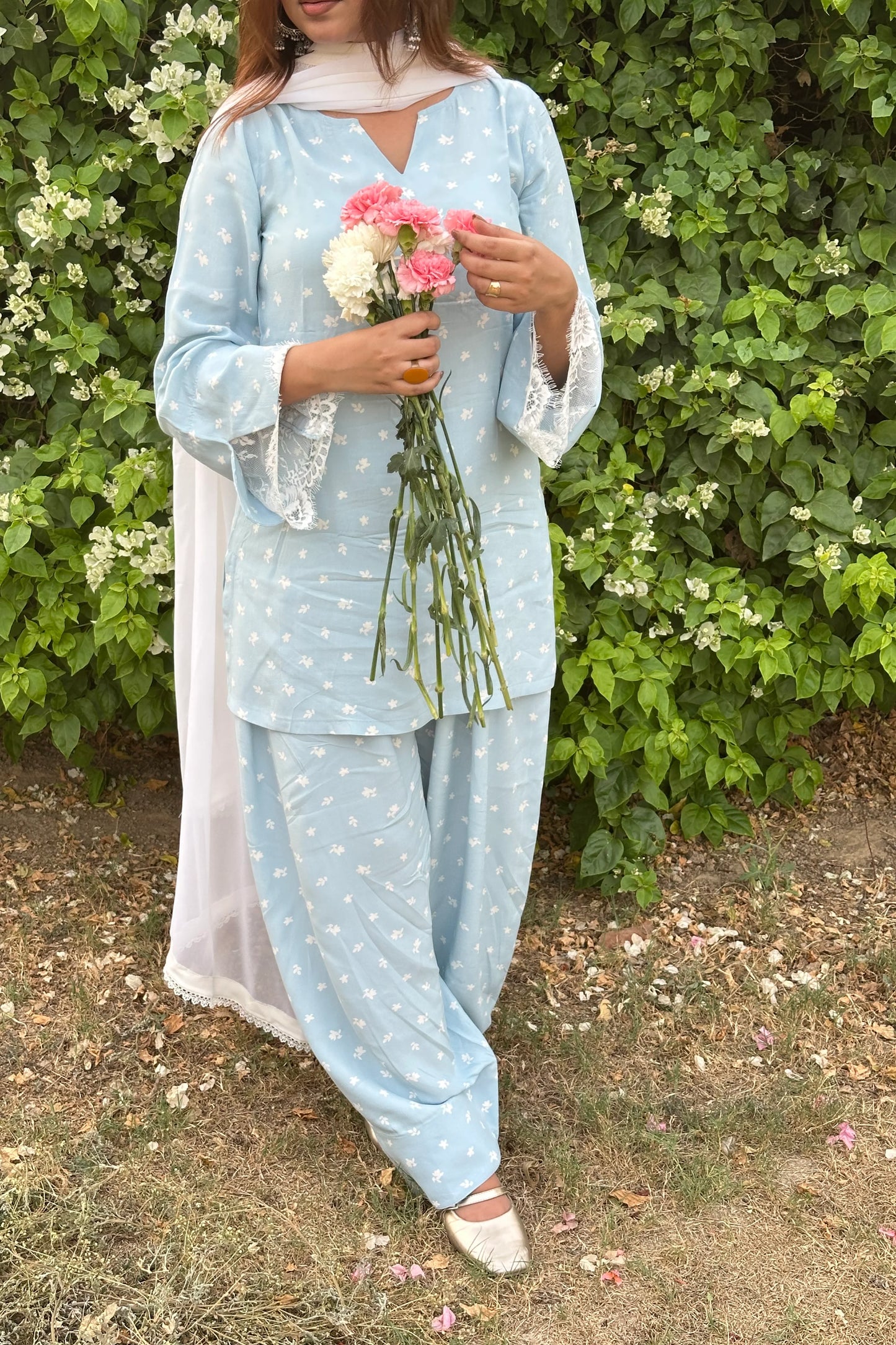 A woman wearing a light blue three-piece salwar kameez set with white star prints, holding a bouquet of pink and white flowers, standing in front of a green leafy wall.
