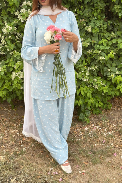 A woman wearing a light blue three-piece salwar kameez set with white star prints, holding a bouquet of pink and white flowers, standing in front of a green leafy wall.
