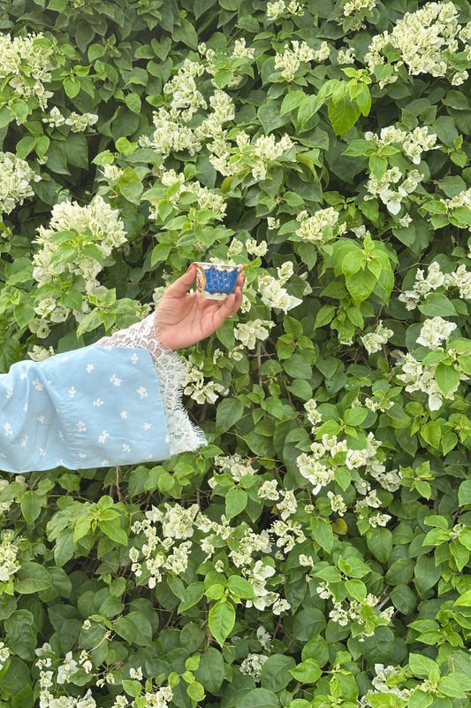 A light blue sleeve with white floral embroidery and lace trim holds a small blue and white patterned cup against a background of green leaves and white flowers.