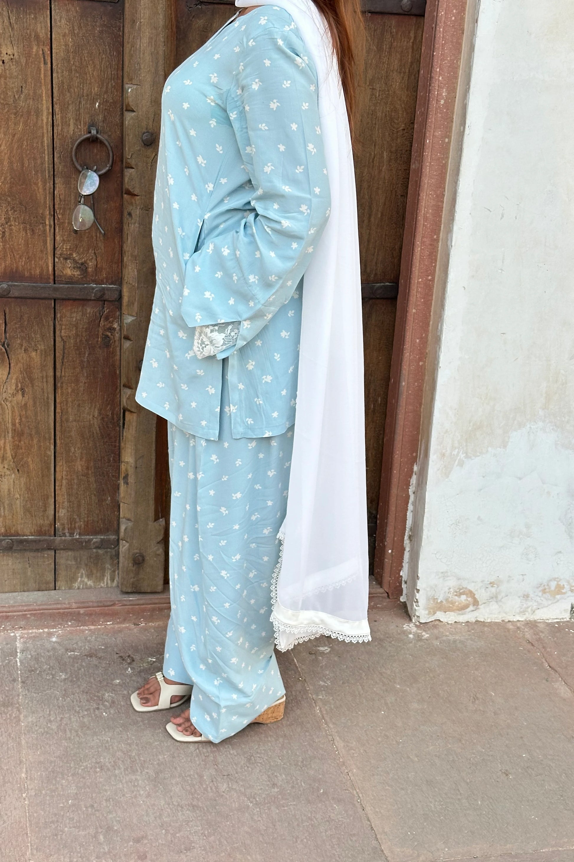A woman wearing a light blue three-piece traditional outfit with small white floral prints, featuring a long-sleeve kurta, matching pants, and a white dupatta, standing in front of a rustic wooden door.