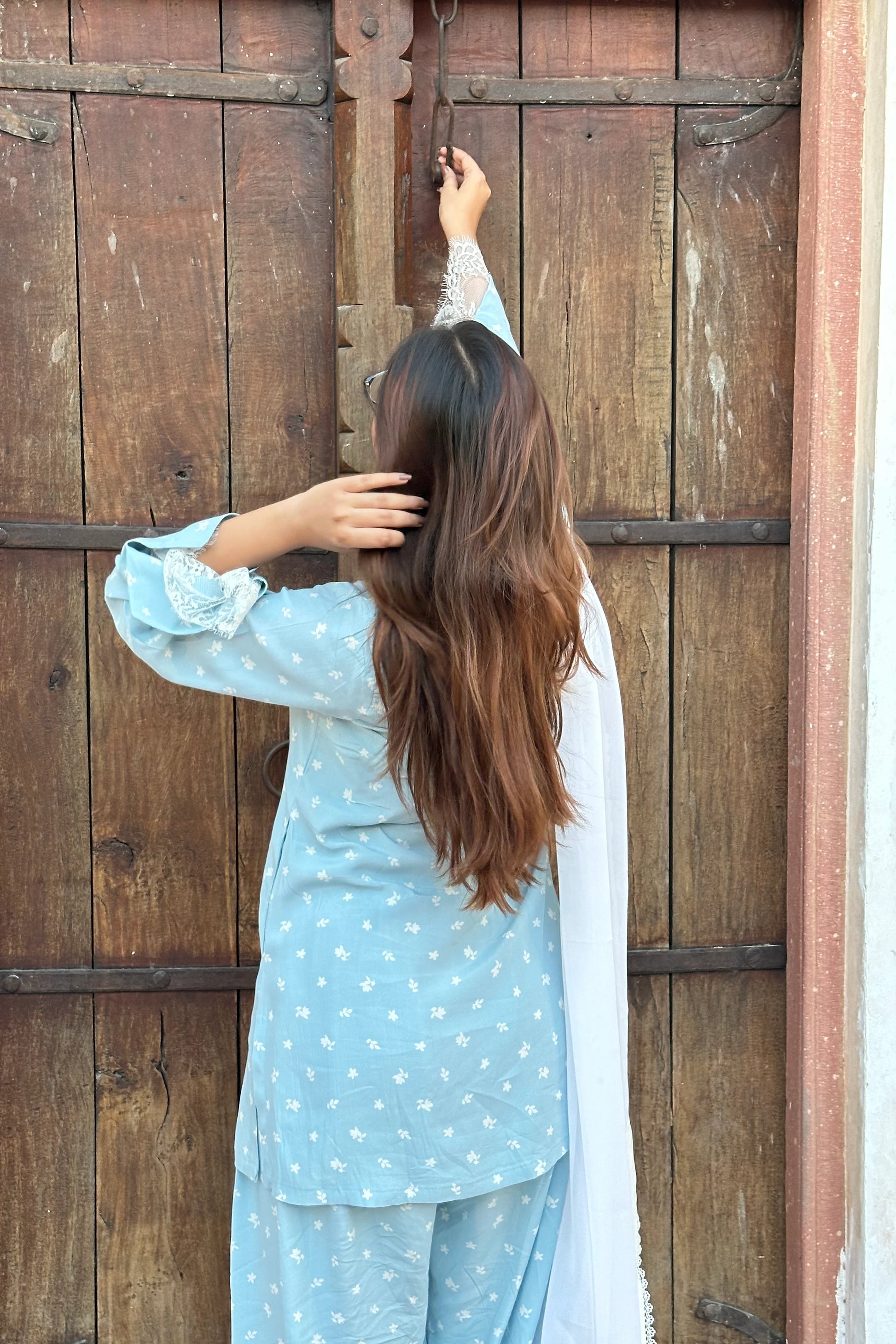 A woman viewed from behind wearing a light blue patterned kurta and matching trousers with a white dupatta, standing in front of a rustic wooden door.