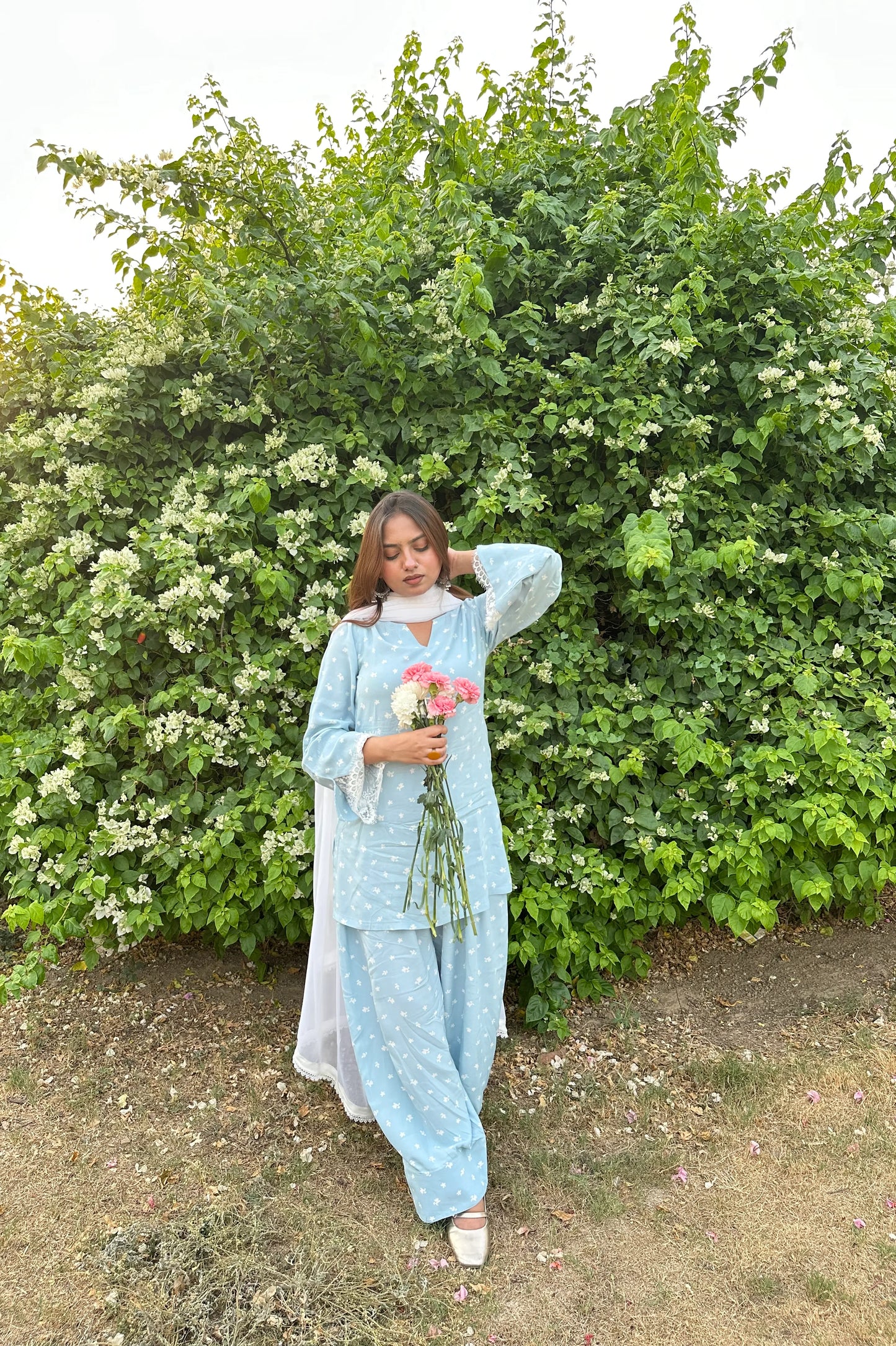 A woman stands outdoors in front of a leafy green hedge, wearing a light blue two-piece outfit with white embroidered patterns and a white inner dress. She is holding a small bouquet of pink and white flowers.