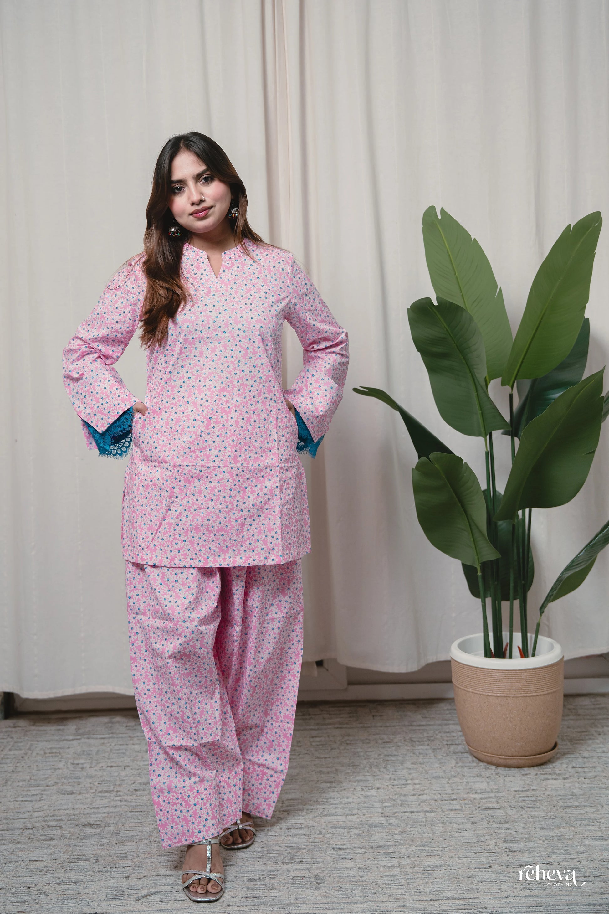 A woman modeling a pink two-piece kurta set with a small white floral pattern, standing in front of a beige curtain next to a potted green plant.