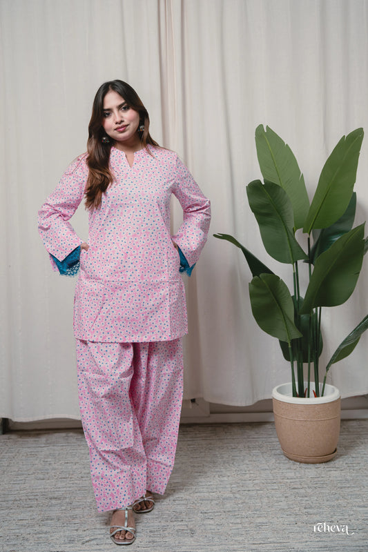 A woman modeling a pink two-piece kurta set with a small white floral pattern, standing in front of a beige curtain next to a potted green plant.