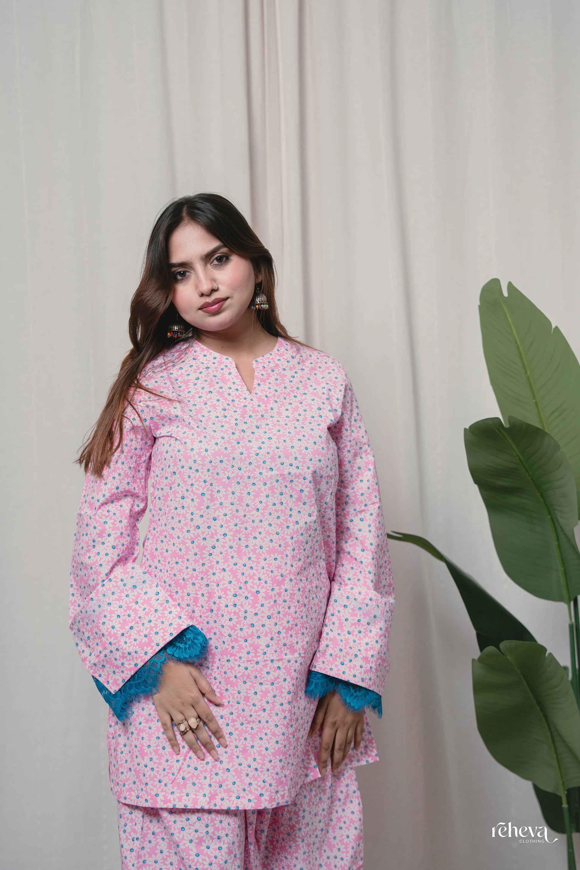 A woman modeling a light pink cotton pajama set with a small blue and green floral print, featuring contrasting blue lace trim on the cuffs. She is standing in front of a neutral cream curtain with a large green plant leaf visible on the right.