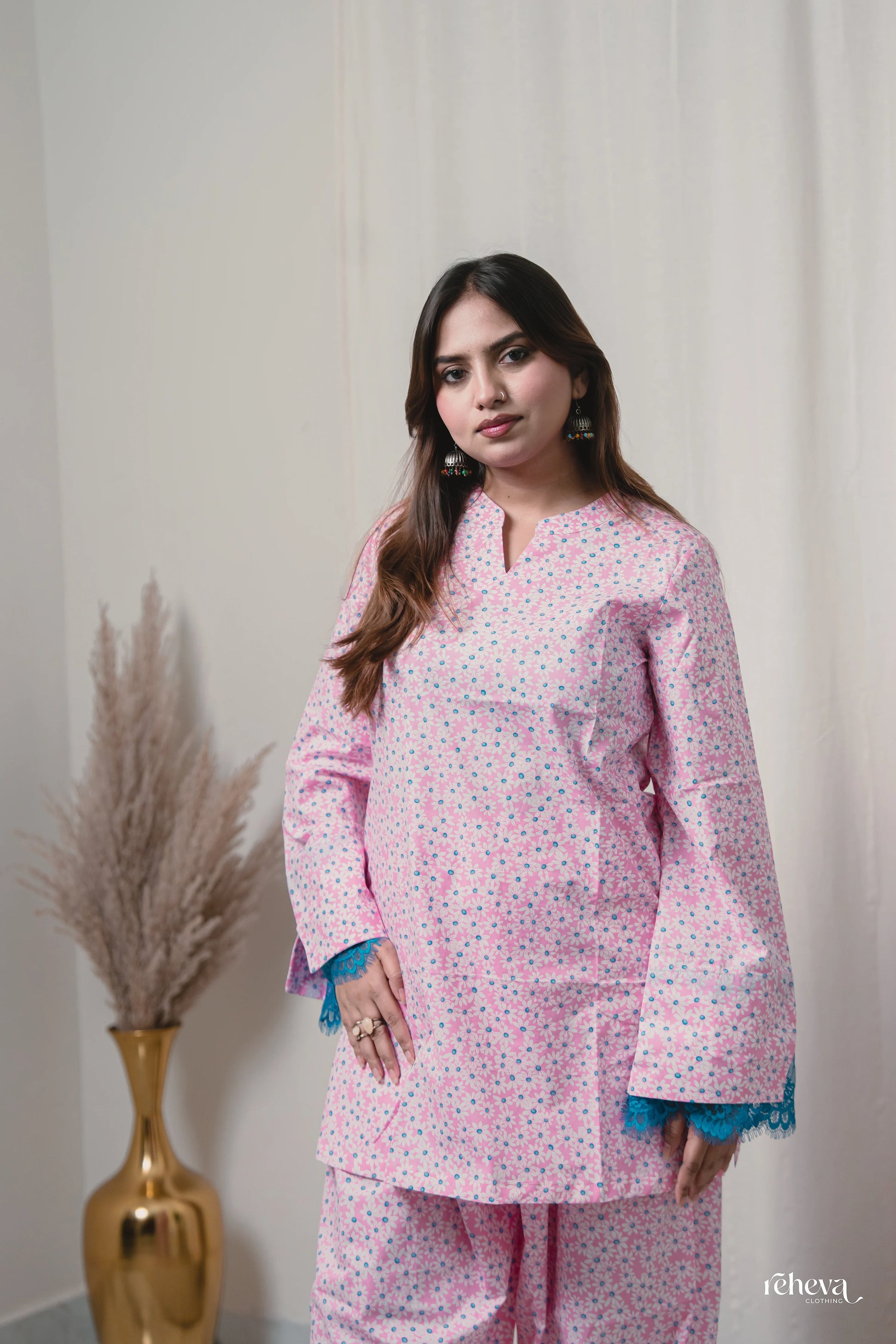 A woman modeling a light pink kurta and matching pants set with a small multicolor floral print, featuring blue tassel trim at the sleeve cuffs, set against a neutral cream background with a gold vase of pampas grass on the left.