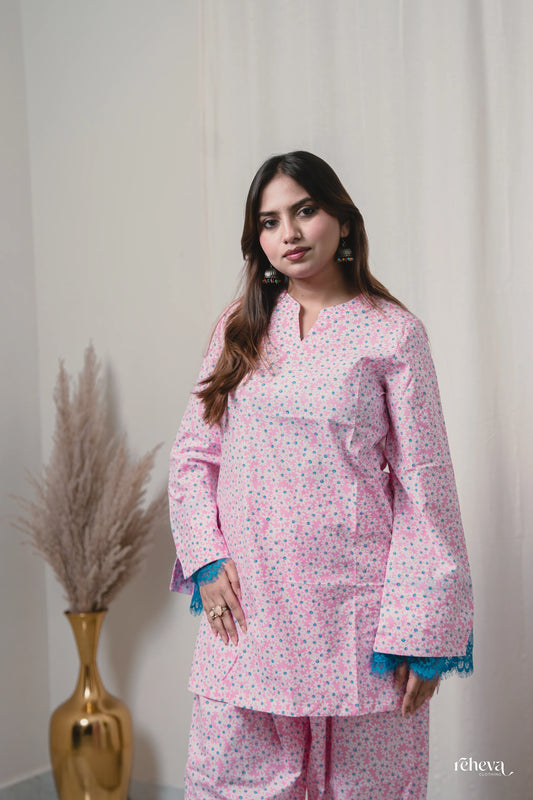 A woman modeling a light pink kurta and matching pants set with a small multicolor floral print, featuring blue tassel trim at the sleeve cuffs, set against a neutral cream background with a gold vase of pampas grass on the left.