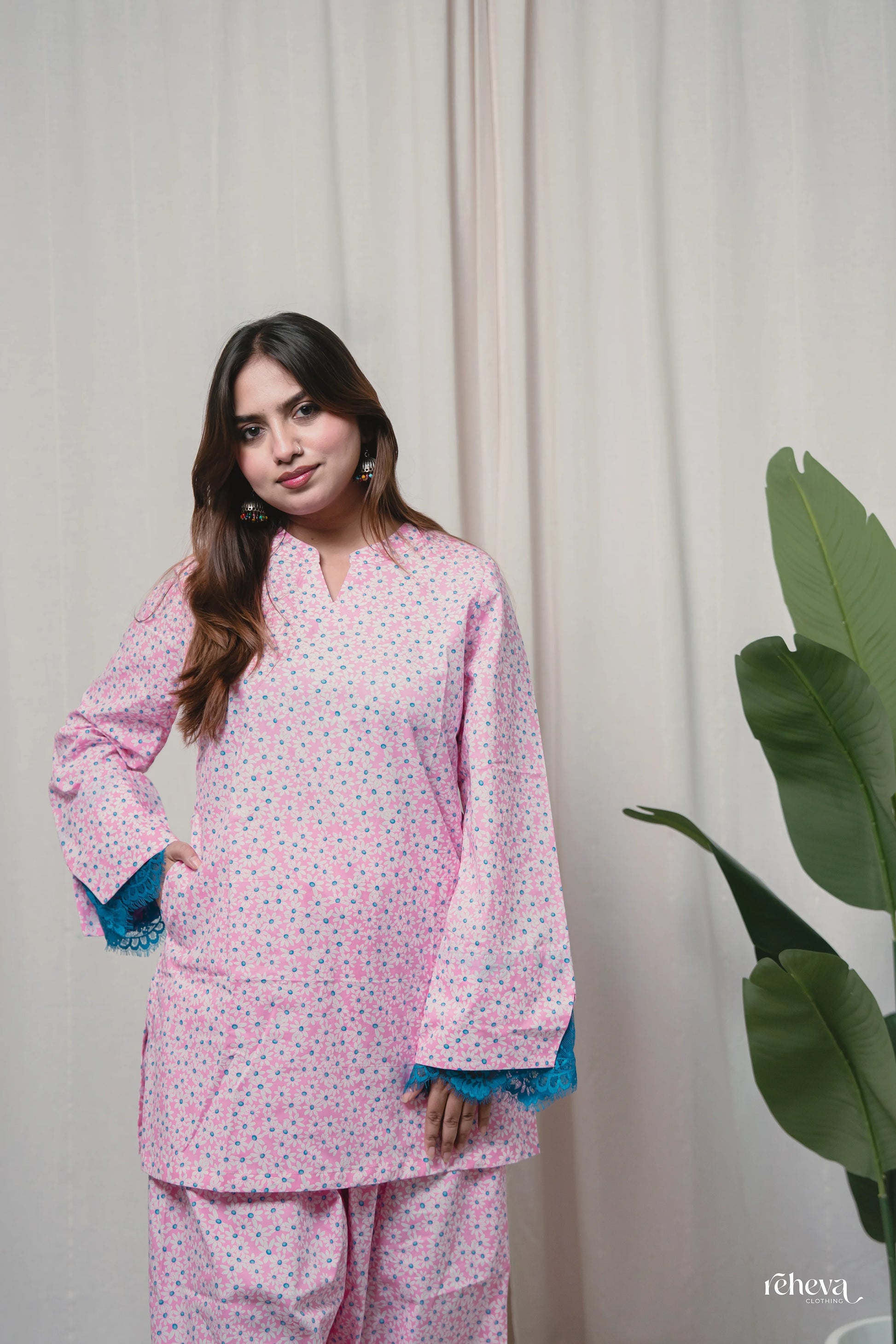 A woman modeling a pink two-piece pajama set with a small multicolor floral print and contrasting blue lace trim on the cuffs, posed against a cream curtain backdrop with a green plant leaf visible on the right.