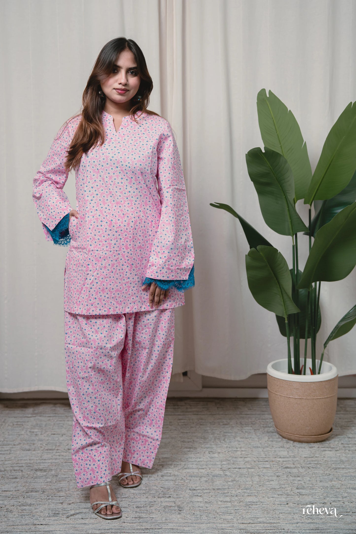 A woman modeling a light pink two-piece pajama set with a small blue and yellow floral pattern, featuring blue-trimmed flared sleeves, standing in front of a neutral curtain next to a potted plant.