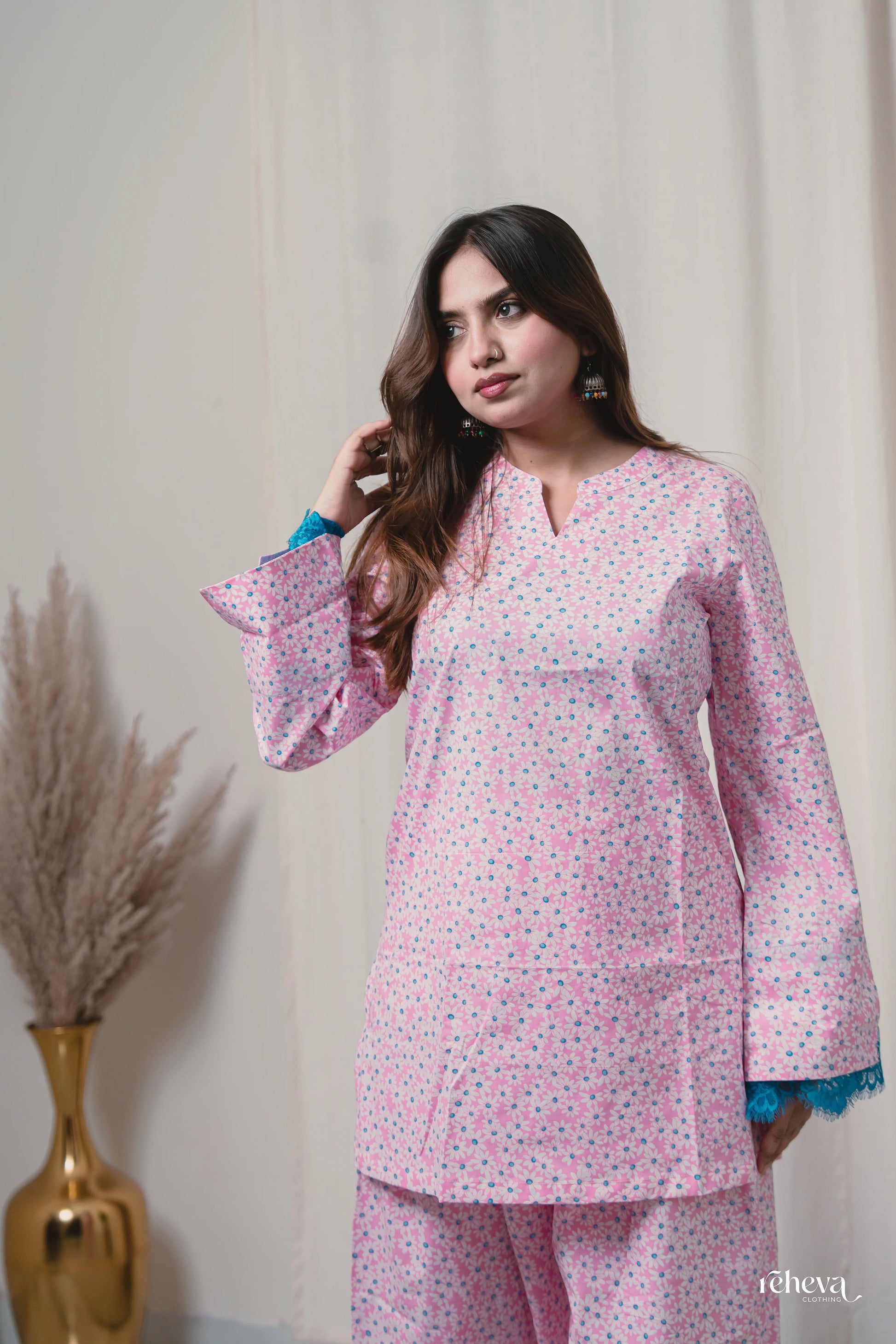 A woman modeling a light pink two-piece kurta set with a small multicolor floral print, featuring blue lace trim on the cuffs, set against a neutral cream background with a decorative gold vase.